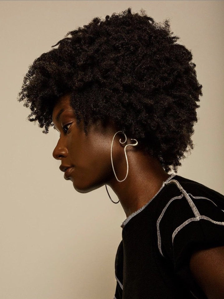 Side profile of model posing against leafy green background, wearing large gold earrings, and a black mesh crop top.